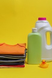 Laundry detergents, stack of baby clothes and rubber duck on yellow background