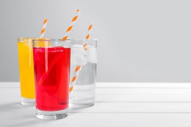 Glasses of different refreshing soda water with ice cubes and straws on white wooden table against light grey background. Space for text