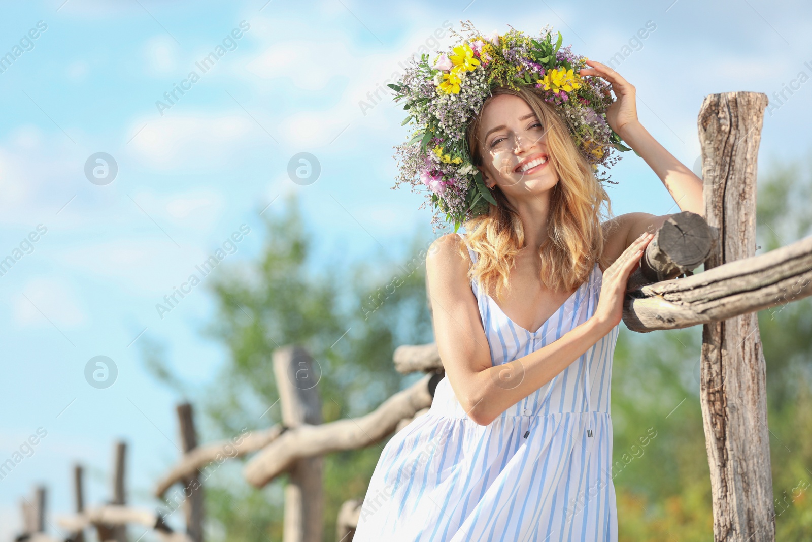 Photo of Young woman wearing wreath made of beautiful flowers near wooden fence on sunny day