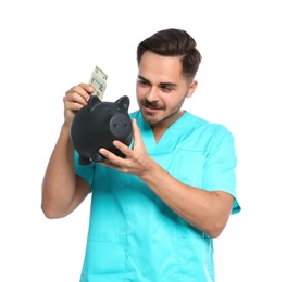 Photo of Portrait of young male doctor putting money into piggy bank on white background