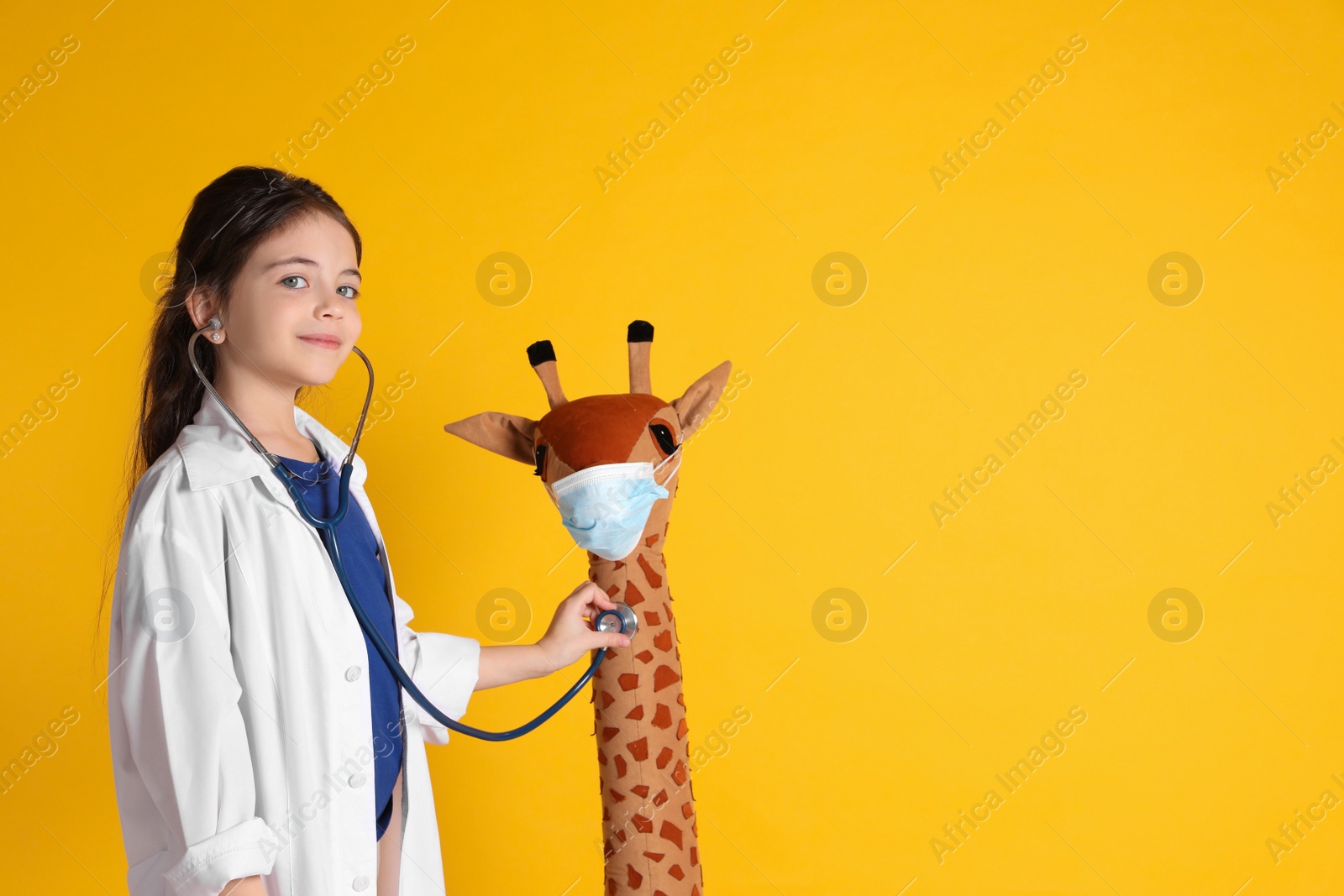 Photo of Little girl playing doctor with toy giraffe on yellow background, space for text. Pediatrician practice