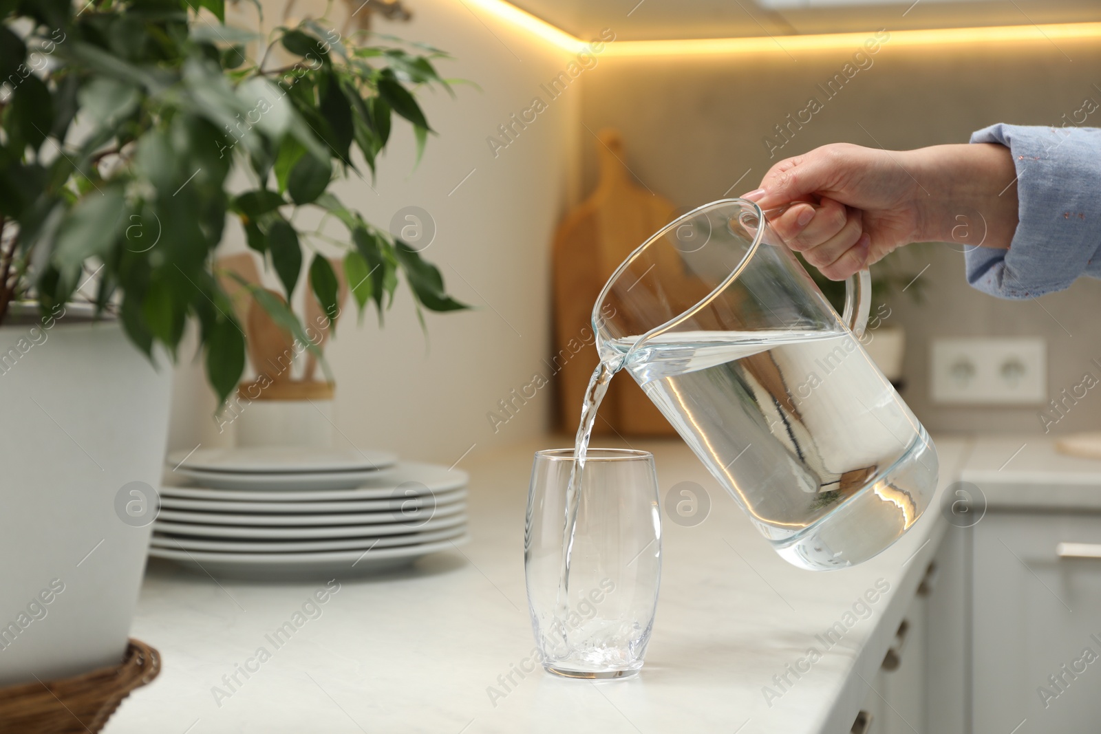 Photo of Woman pouring water from jug into glass at white table in kitchen, closeup