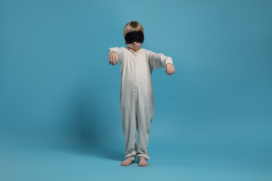 Boy in pajamas and sleep mask sleepwalking on light blue background