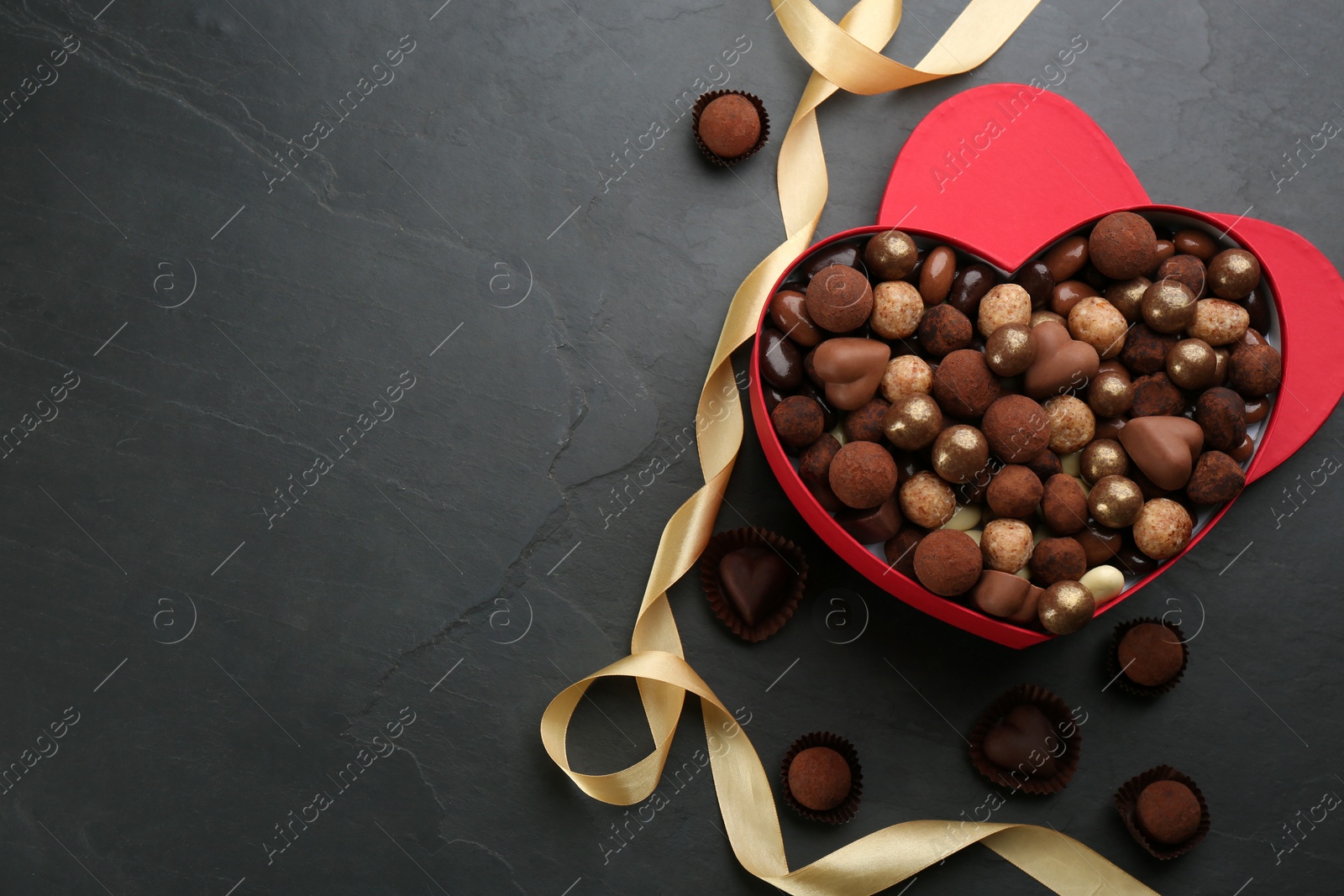 Photo of Different delicious chocolate candies in heart shaped box and ribbon on black table, flat lay. Space for text