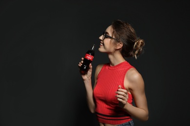 Photo of MYKOLAIV, UKRAINE - NOVEMBER 28, 2018: Young woman with bottle of Coca-Cola on dark background, space for text
