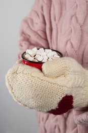 Photo of Woman in knitted mittens holding cup of delicious hot chocolate with marshmallows on light background, closeup