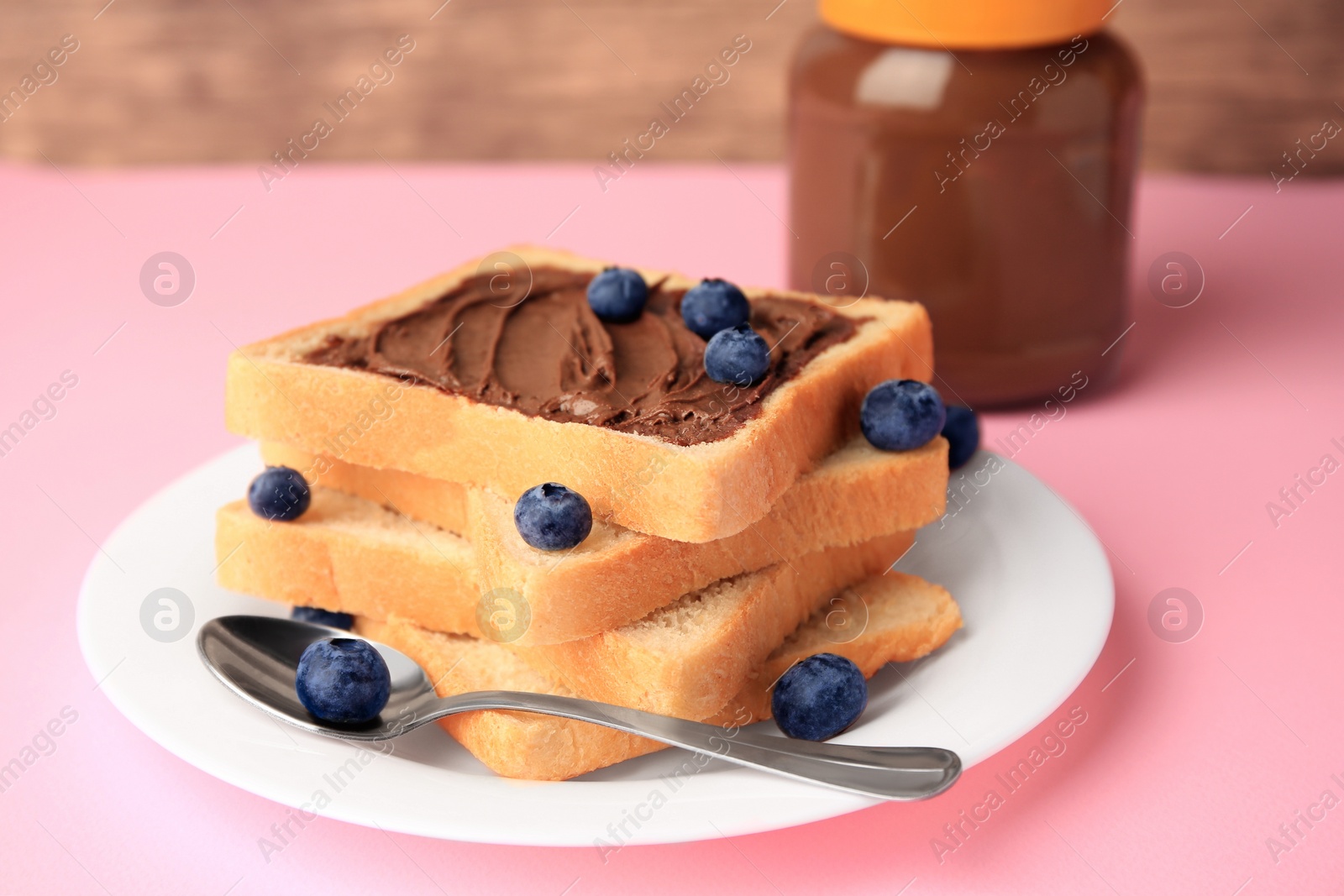 Photo of Tasty toast with chocolate paste and blueberries near jar on pink table, closeup