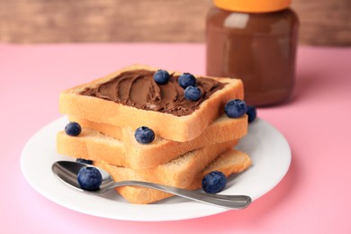 Photo of Tasty toast with chocolate paste and blueberries near jar on pink table, closeup