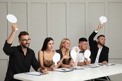 Photo of Panel of judges with different emotions holding blank score signs at table against light wall. Space for text