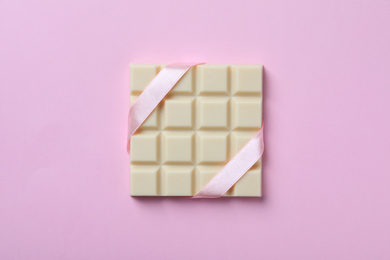 Photo of Tasty white chocolate bar with ribbon on pink background, top view