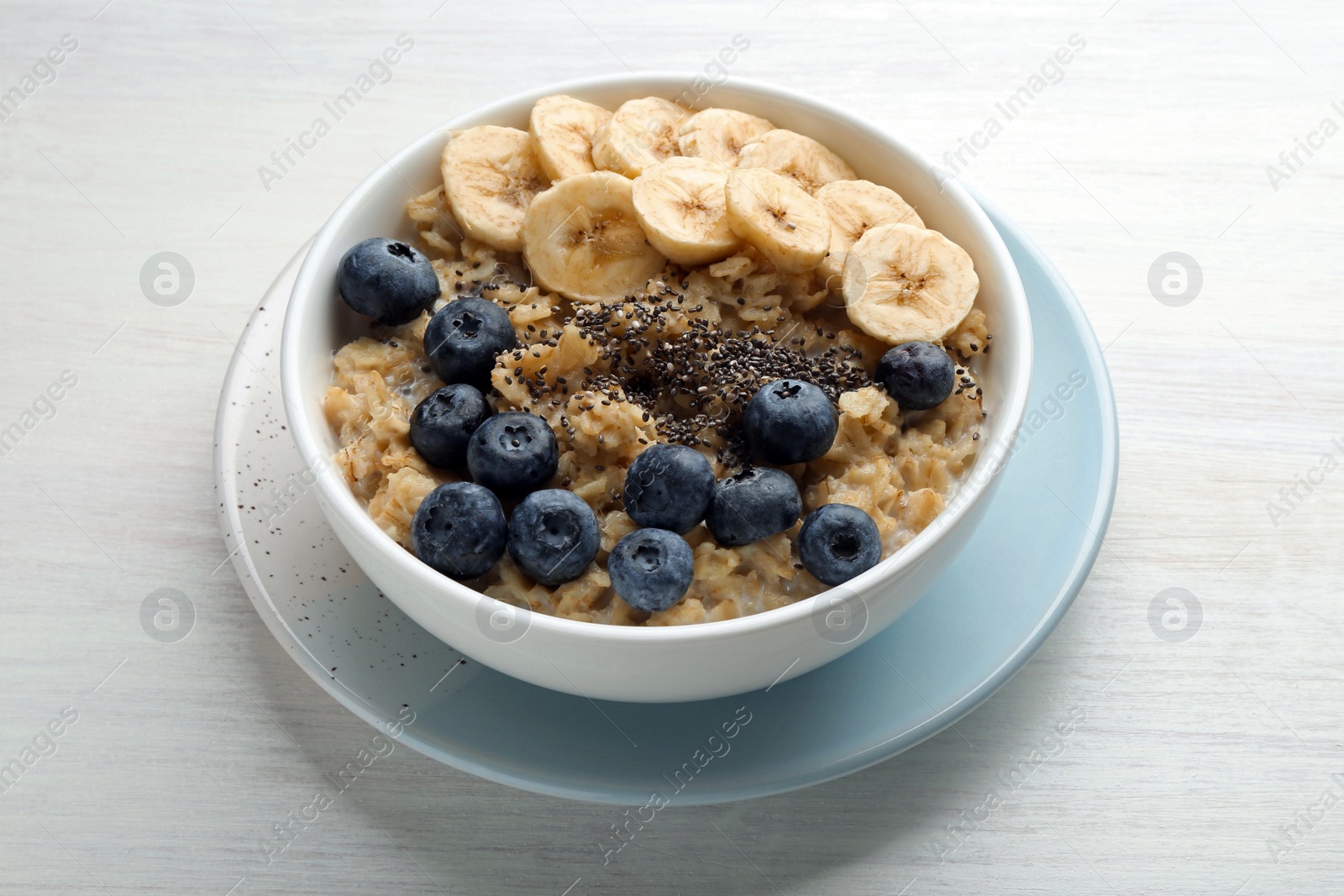 Photo of Tasty oatmeal with banana, blueberries and chia seeds served in bowl on white wooden table