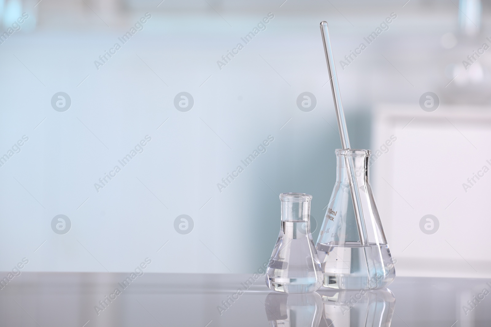 Photo of Laboratory analysis. Glass flasks and stirring rod on white table against blurred background, space for text