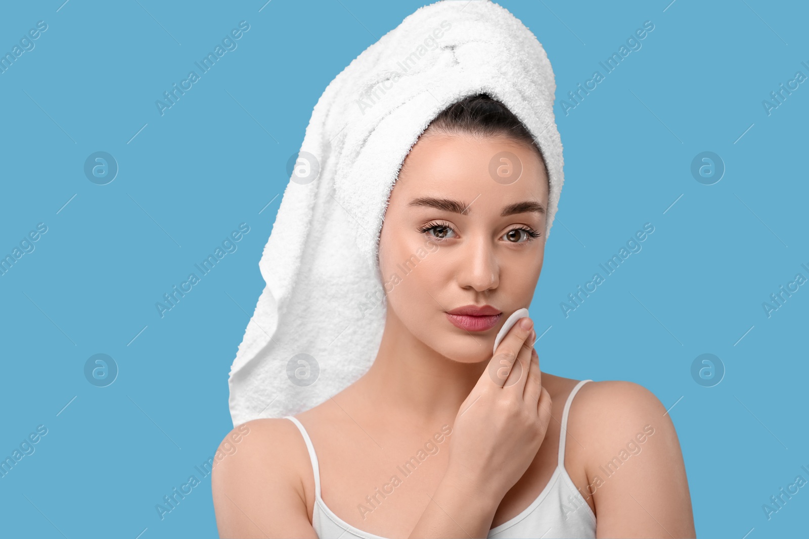 Photo of Beautiful woman in terry towel removing makeup with cotton pad on light blue background