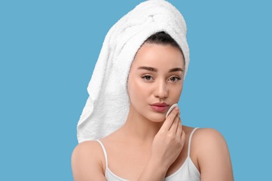 Beautiful woman in terry towel removing makeup with cotton pad on light blue background