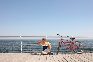 Attractive man with laptop and bike near sea on sunny day, space for text