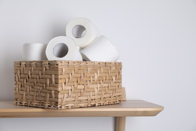 Toilet paper rolls in wicker basket on wooden table near white wall, space for text