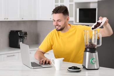 Photo of Happy man adding berries into blender while watching cooking online course in kitchen. Time for hobby