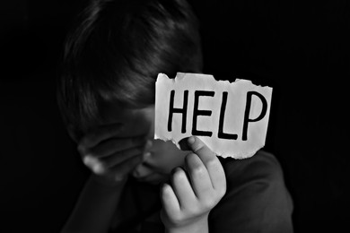 Little boy holding piece of paper with word Help against black background, focus on hand. Domestic violence concept