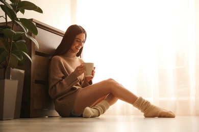 Photo of Woman with cup of drink sitting on warm floor at home. Heating system