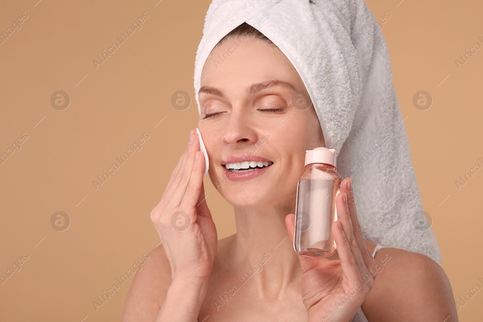 Photo of Beautiful woman in terry towel removing makeup with cotton pad on beige background, space for text