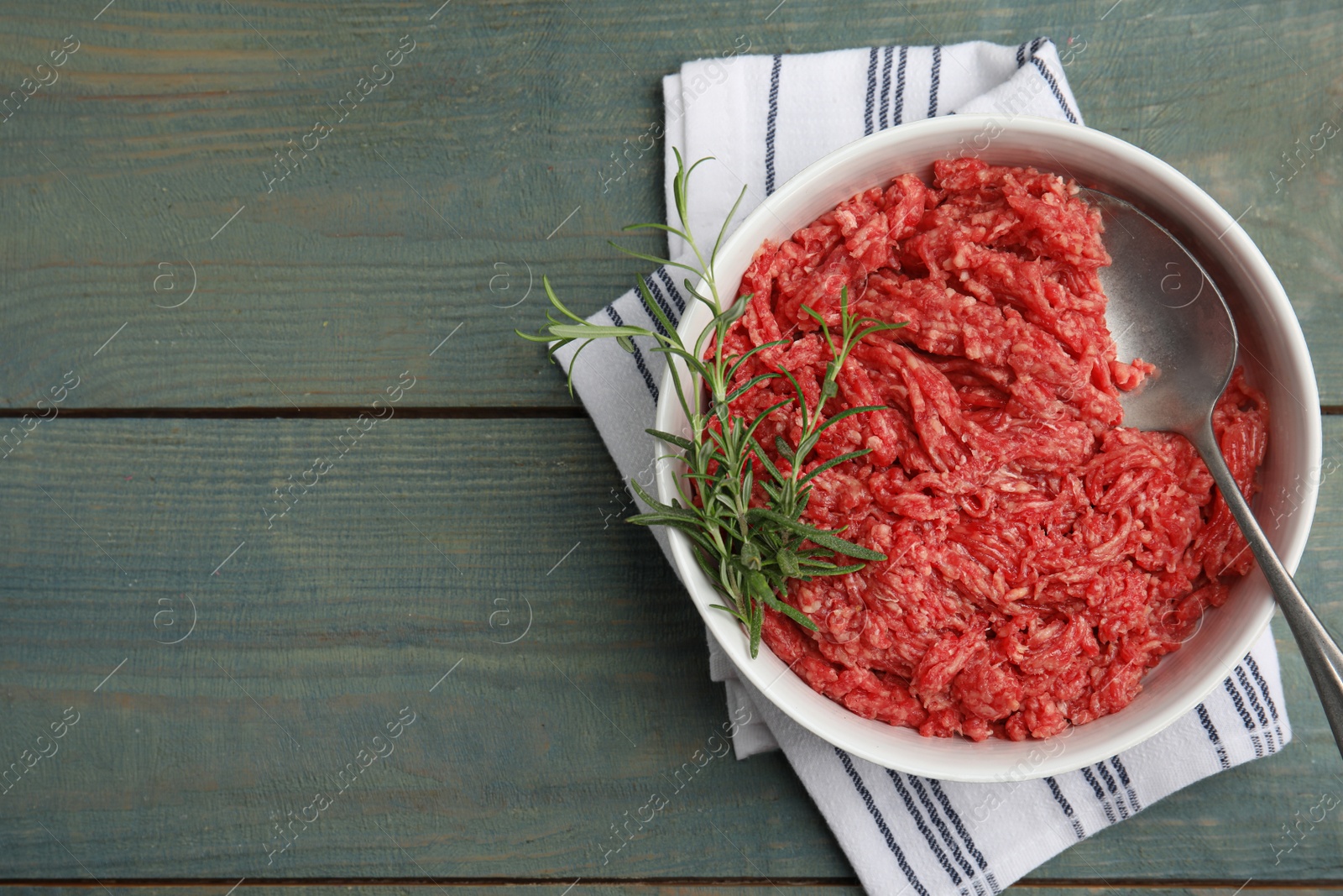 Photo of Fresh minced meat and spoon in bowl on light blue wooden table, top view. Space for text