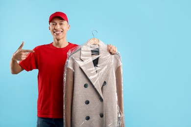 Dry-cleaning delivery. Happy courier holding coat in plastic bag on light blue background, space for text