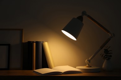 Photo of Stylish modern desk lamp, books and plant on wooden table in dark room