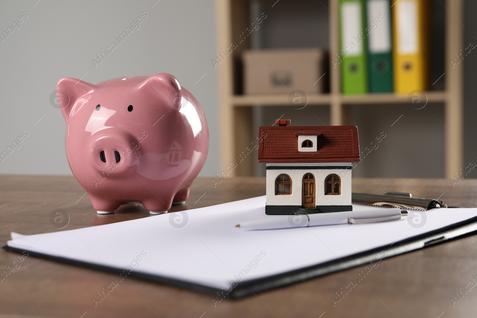 Photo of House model, piggy bank, clipboard and pen on wooden table indoors