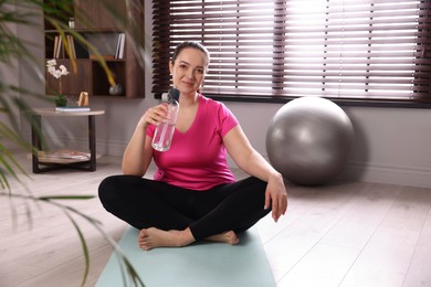 Photo of Overweight woman with bottle of water on yoga mat at home
