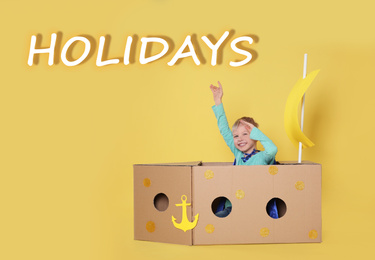 Image of School holidays. Cute little child playing with cardboard ship near yellow wall