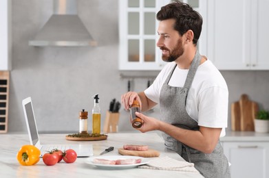 Photo of Man making dinner while watching online cooking course via laptop in kitchen