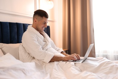 Photo of Handsome man wearing bathrobe with laptop on bed in hotel room