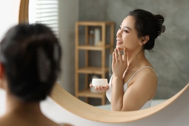 Photo of Happy woman applying face cream near mirror at home