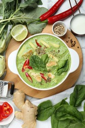 Saucepan with delicious green curry chicken soup and different ingredients on white marble table, flat lay