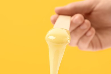 Photo of Woman holding spatula with hot depilatory wax on yellow background, closeup