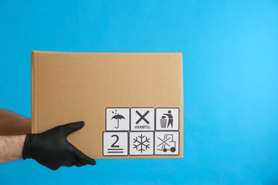 Photo of Courier holding cardboard box with different packaging symbols on blue background, closeup. Parcel delivery
