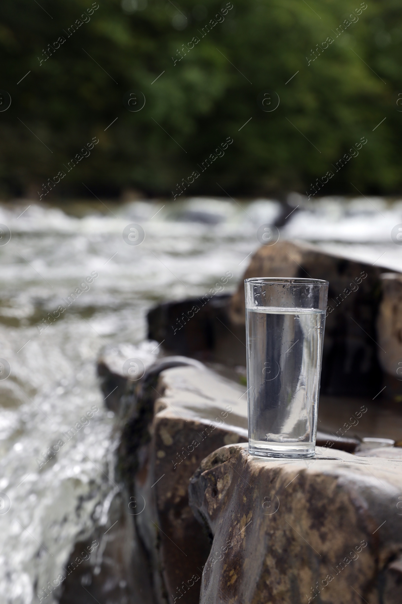 Photo of Glass of fresh water on stone near river, space for text