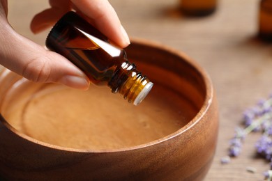 Woman dripping lavender essential oil from bottle into wooden bowl at table, closeup