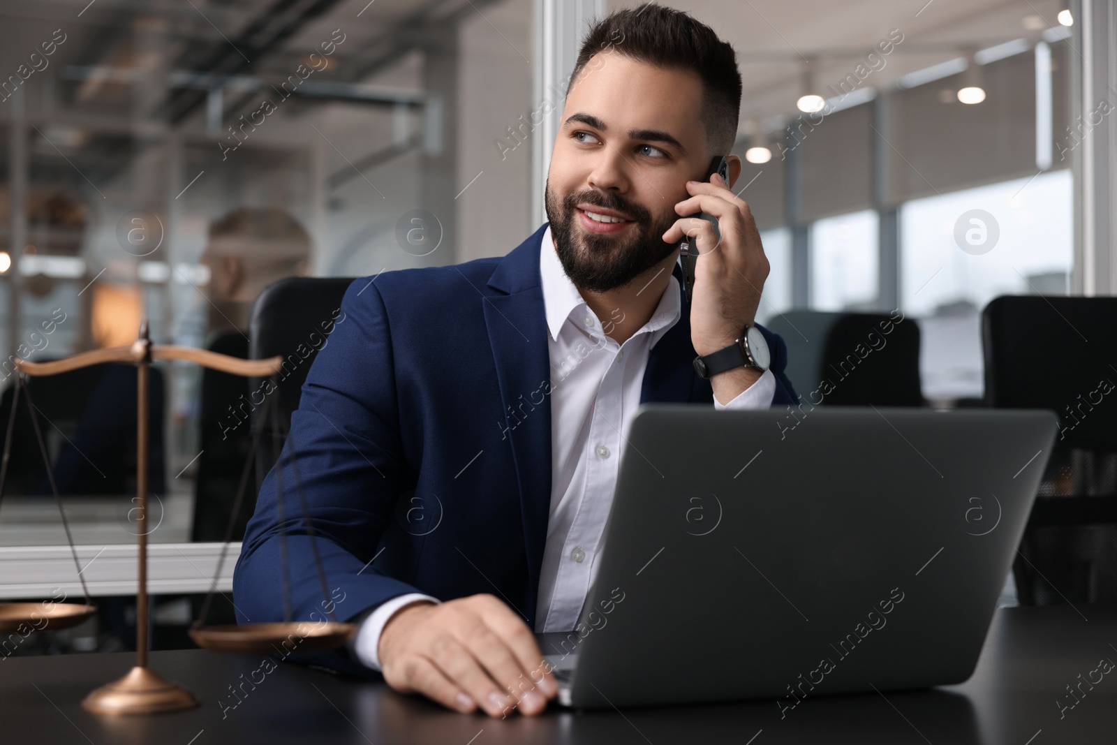 Photo of Smiling lawyer talking on smartphone near laptop at table in office