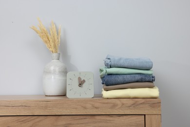 Different folded clothes, analog clock and dry plants on wooden chest of drawers near grey wall