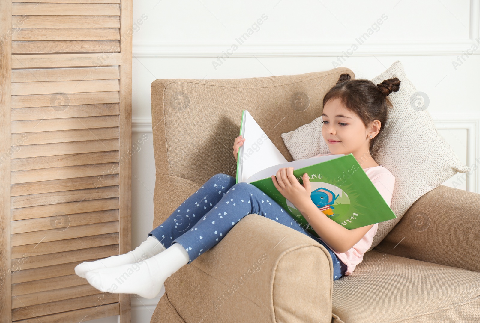 Photo of Little girl reading book in armchair at home