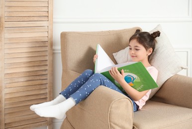 Photo of Little girl reading book in armchair at home