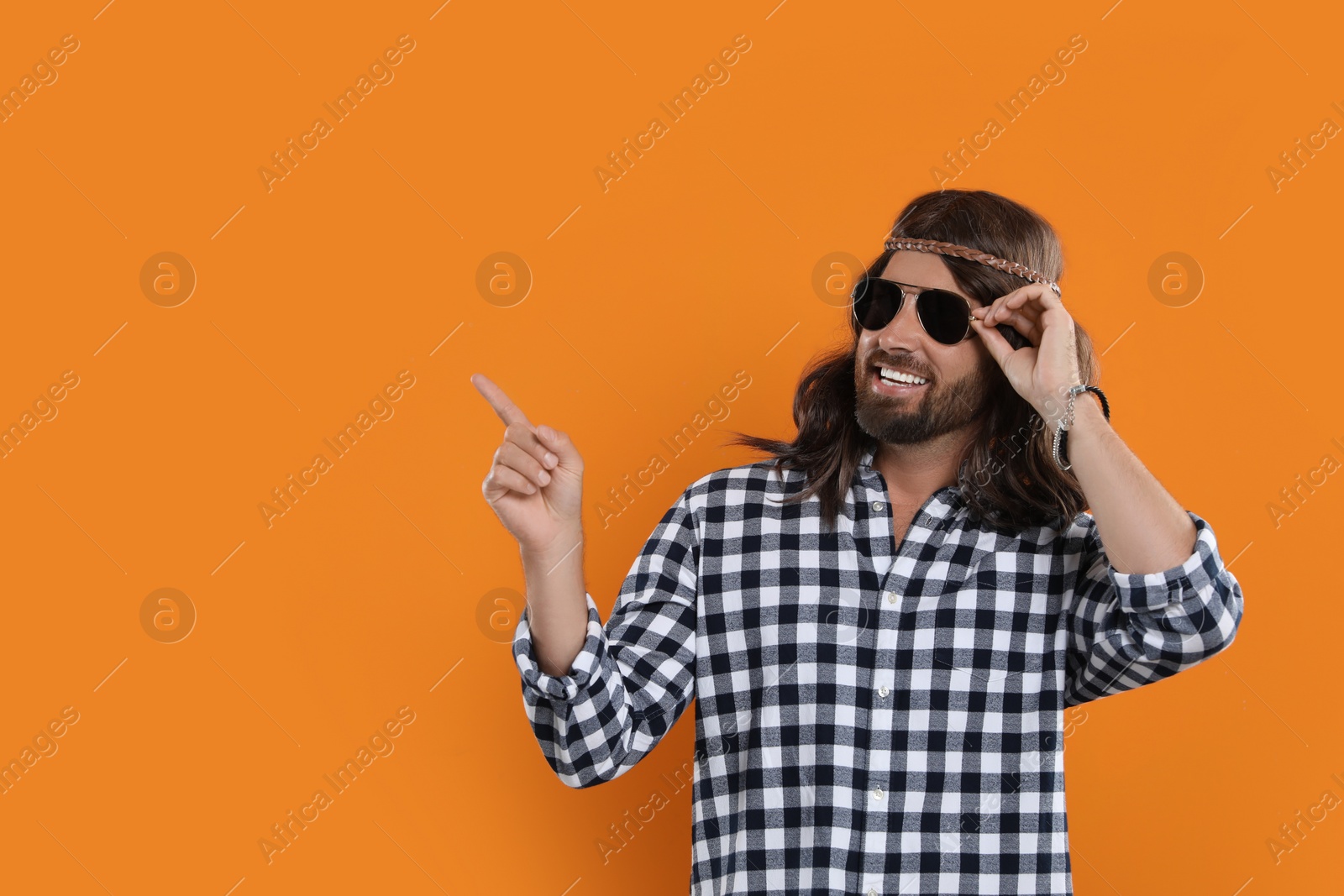 Photo of Hippie man in sunglasses pointing at something on orange background, space for text