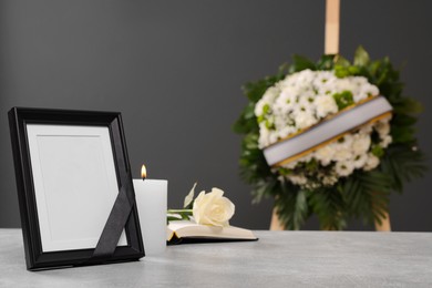 Photo frame with black ribbon, rose, burning candle on light table and wreath of flowers near grey wall indoors, space for text. Funeral attributes
