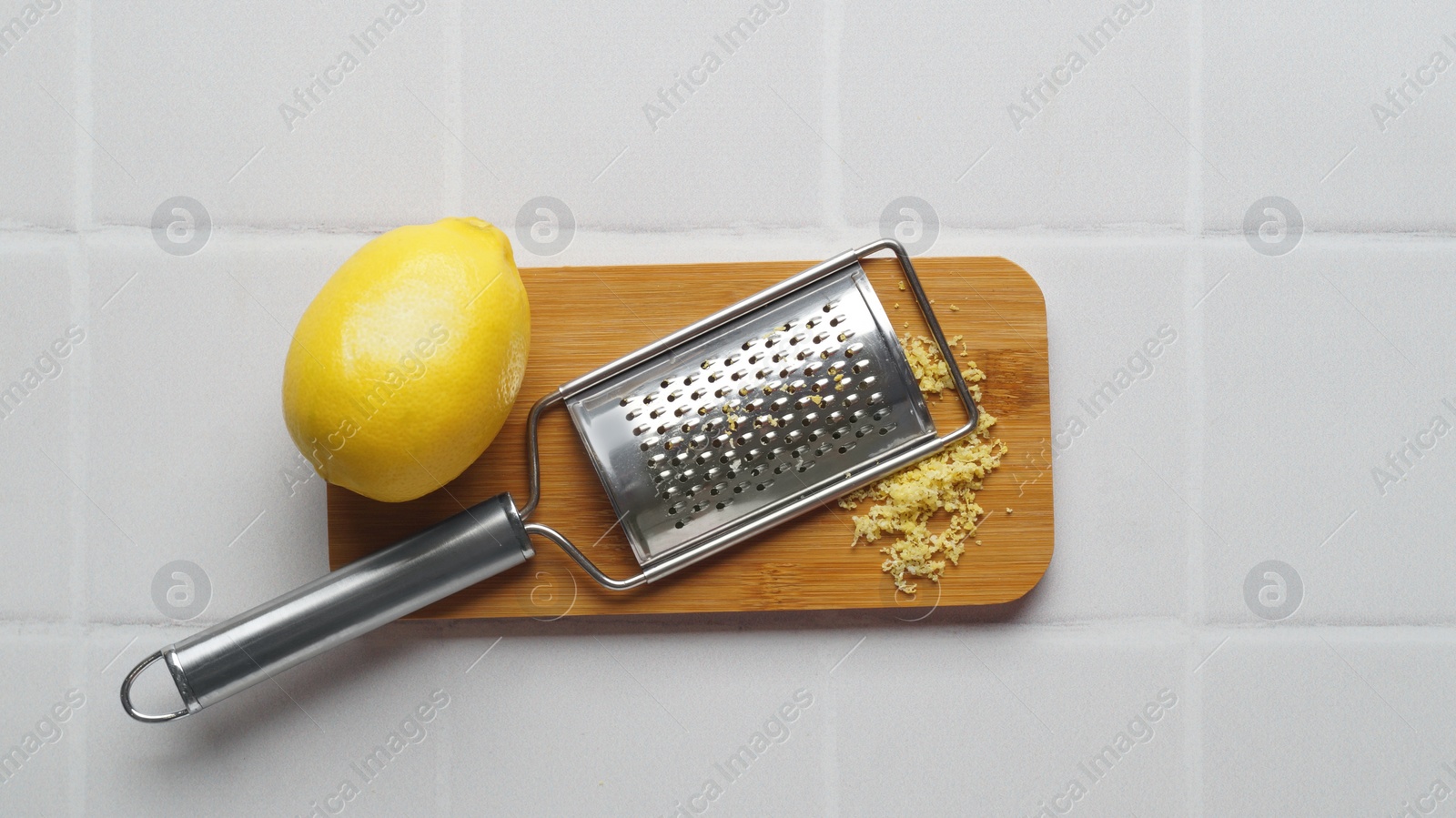 Photo of Wooden board with grater and fresh lemon zest on white tiled table, flat lay