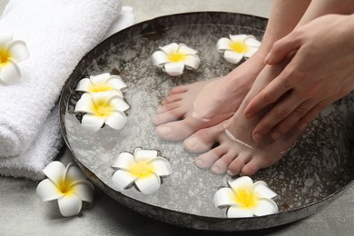 Woman soaking her feet in bowl with water and flowers on light grey floor, closeup. Spa treatment