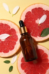 Photo of Bottle of cosmetic serum, grapefruit slices and green leaves on wet yellow background, flat lay