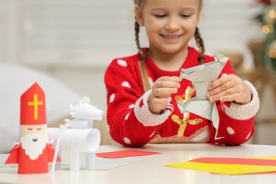 Photo of Cute little girl making paper toy for Saint Nicholas day at home, focus on hands