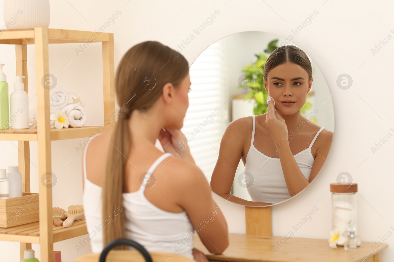 Photo of Beautiful woman removing makeup with cotton pad near mirror at home