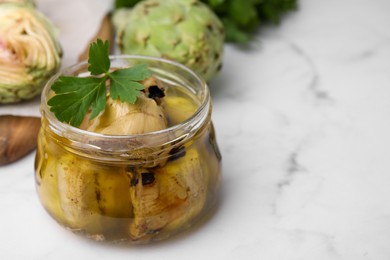 Photo of Jar of delicious artichokes pickled in olive oil on white marble table, closeup. Space for text